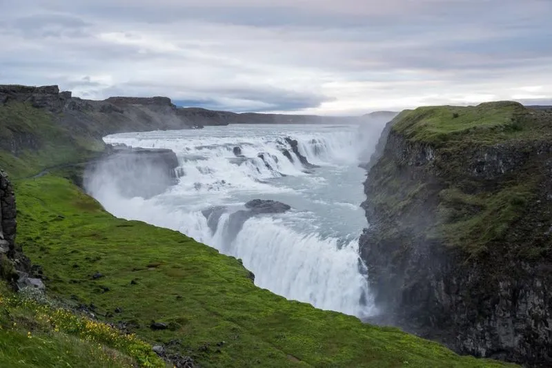 Islanda: Ampia cascata tra pareti rocciose e prati verdi sotto cielo nuvoloso