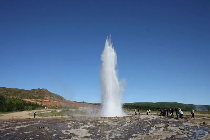 Islanda: Geyser in eruzione con getto d'acqua alto circondato da visitatori e terreno roccioso