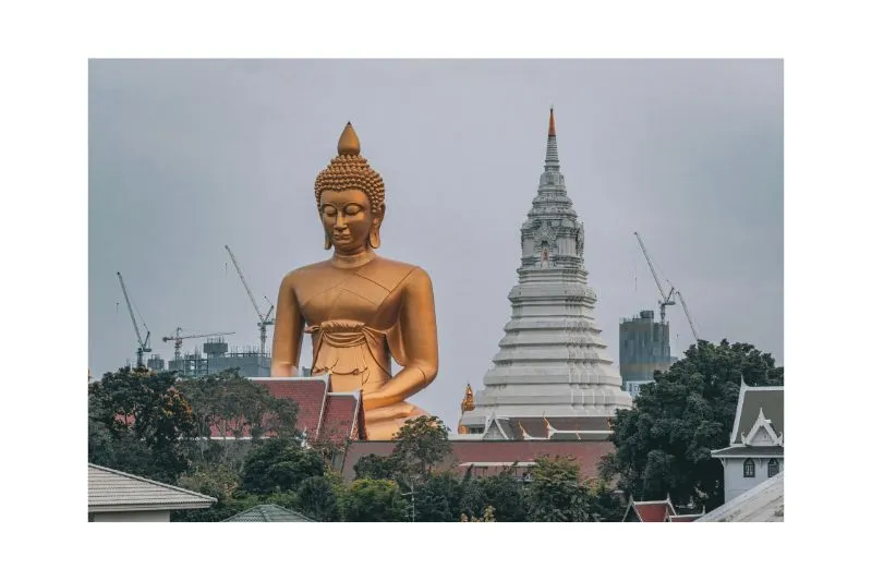 Grande statua dorata del Buddha a Bangkok con stupa bianca di un tempio sullo sfondo e skyline urbano.