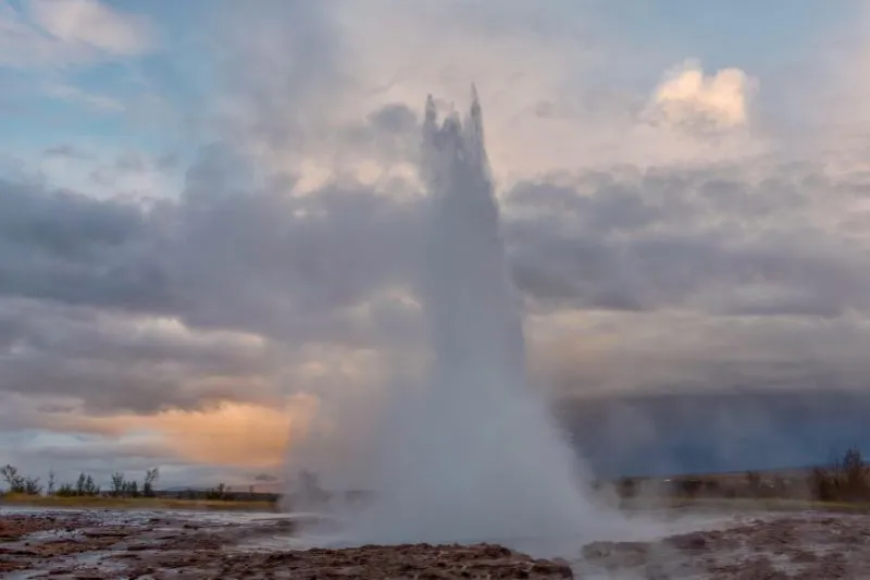 Geyser in eruzione con getto d’acqua e vapore sotto cielo nuvoloso